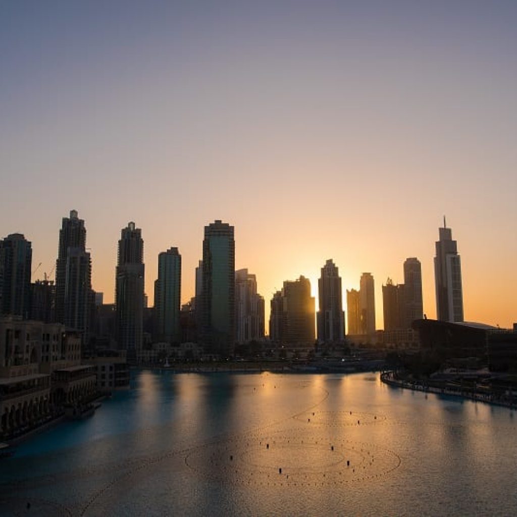 Muchos rascacielos situados tras el mar, reflejándose en el agua, con la puesta de Sol de fondo. Viajar a Dubai.