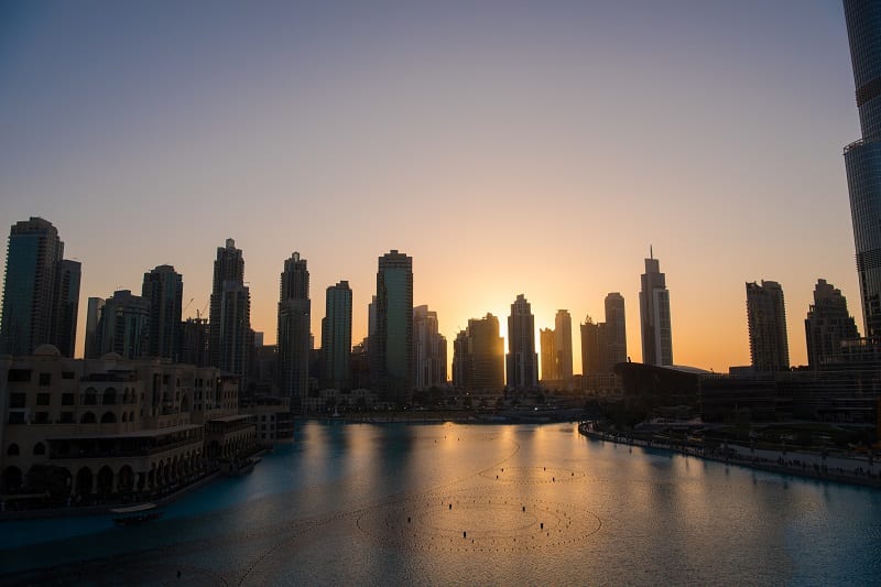 Muchos rascacielos situados tras el mar, reflejándose en el agua, con la puesta de Sol de fondo. Viajar a Dubai.
