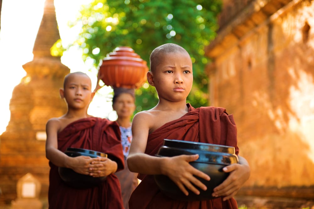 Un niño en primer plano porta un cuenco y viste una toga holgada. De fondo, se ven a un niño haciendo lo mismo y a una mujer con un gran cuenco sobre su cabeza. Viaje Myanmar.