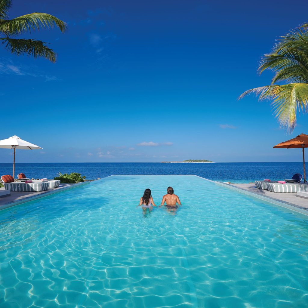 Época viajar Maldivas. Dos personas en una piscina, mirando hacia al mar. Se muestran a las personas de espaldas, y al océano de fondo.